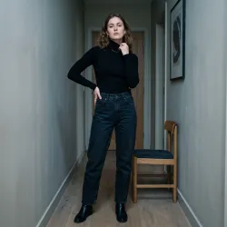 Create a product model image of a black fitted turtleneck top. The model is a beautiful adult young white woman wearing dark relaxed jeans. She is standing in a narrow hallway, one hand on her hip and the other lightly touching her collar, with a strong quiet expression. The background is a simple interior wall with muted paint and a single chair. The lighting is cool and low-key, with a minimal modern mood.