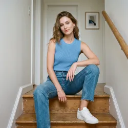 Create a product model image of a sky-blue sleeveless knit top. The model is a beautiful adult young white woman wearing straight-leg vintage jeans. She is sitting on a simple staircase, one knee raised and one arm draped over it, with a soft playful expression. The background is an ordinary hallway with off-white walls and minimal decor. The lighting is cool and soft, with a quiet modern everyday feel.